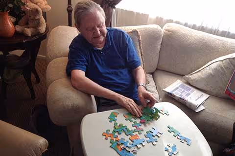 An elderly man sitting on a beige couch in a living room, working on assembling a colorful jigsaw puzzle on a small white table in front of him. There is a newspaper and a pillow on the couch beside him, and a small table with a teddy bear and a vase in the background.