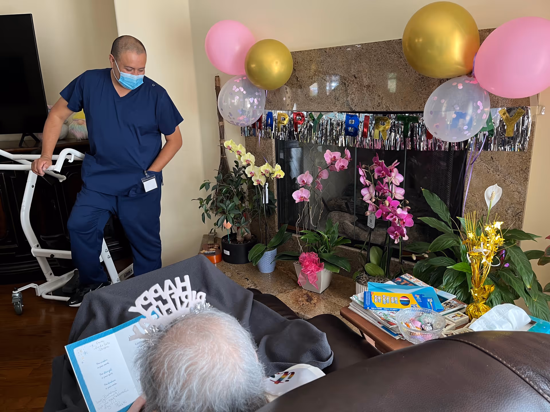 A man in blue scrubs and a face mask stands near a white assistive device in a living room decorated for a birthday celebration with balloons, orchids, and a 'Happy Birthday' banner on the fireplace. An elderly person with gray hair wearing a 'Happy Birthday' tiara is seated in the foreground, holding a birthday card.