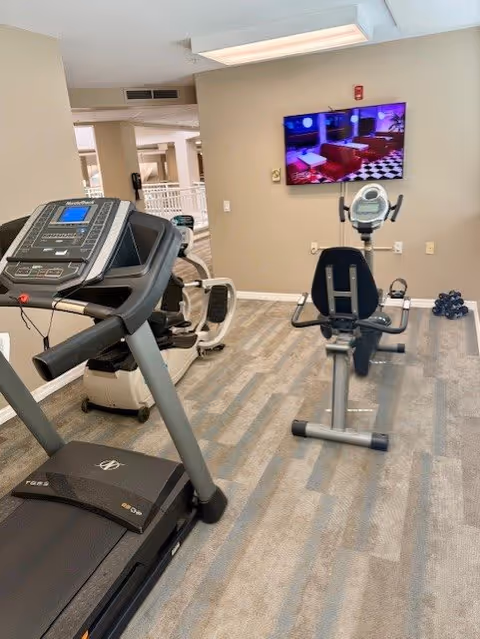 Indoor fitness room with a treadmill and a recumbent exercise bike on a carpeted floor. A flat-screen TV is mounted on the beige wall in front of the exercise equipment, displaying a colorful image. The room has neutral-colored walls and an open doorway leading to another area.
