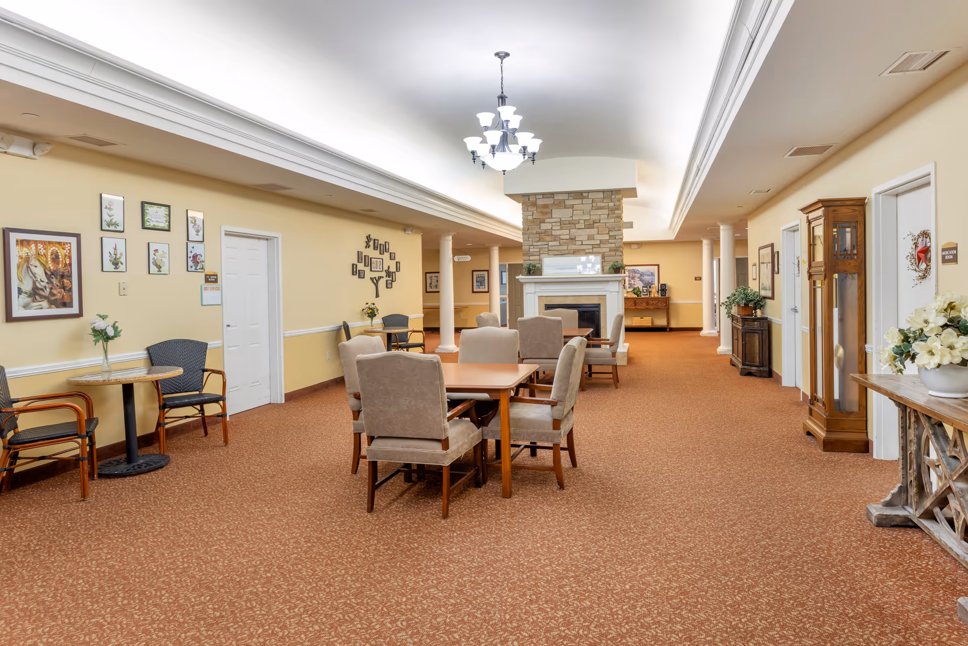Spacious common room with tables and upholstered chairs centered around a stone fireplace in a senior living facility.