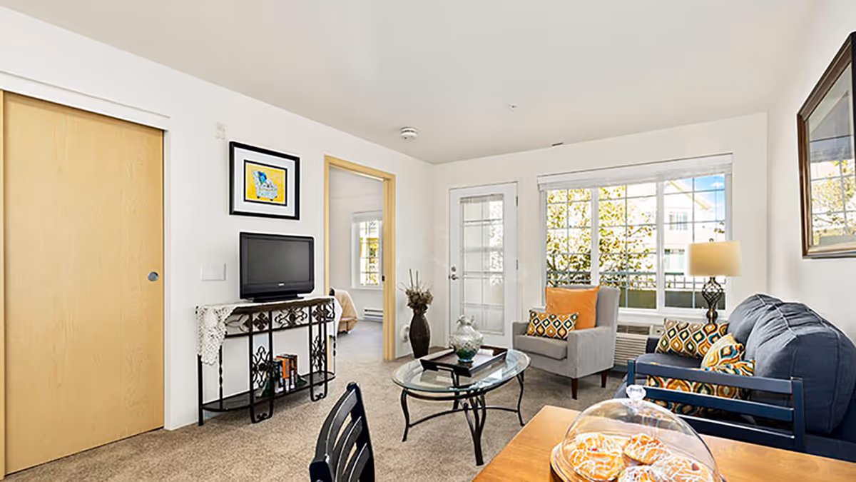 Bright furnished living room with a sofa, armchair, glass coffee table, TV on a console, and a large window and door letting in natural light.