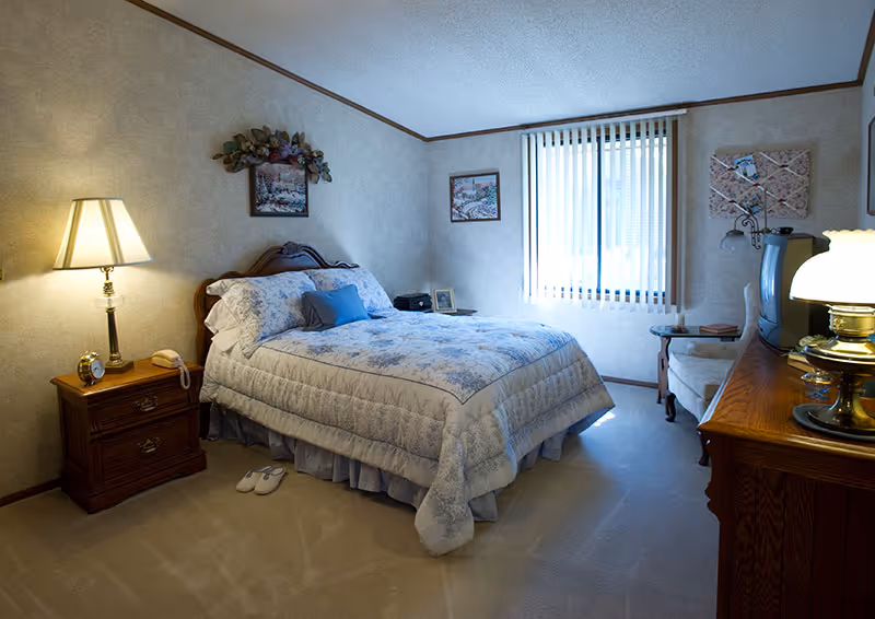A cozy bedroom with a double bed covered in a floral comforter and pillows. There is a wooden nightstand with a lamp, alarm clock, and telephone on the left side of the bed. A pair of slippers is placed on the carpeted floor next to the nightstand. The room has beige walls with framed artwork and a window with vertical blinds letting in natural light. On the right side, there is a wooden dresser with a lamp and an old-style TV, and a cushioned chair next to the window.