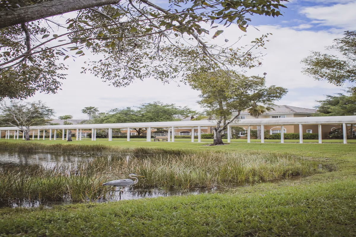 A serene outdoor scene at Aston Gardens At Pelican Marsh featuring a small pond with tall grasses and a heron standing in the water. Surrounding the pond is a well-maintained grassy area with trees and a long covered walkway supported by white columns. In the background, there is a beige building partially visible under a partly cloudy sky.