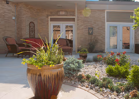 Outdoor patio area with a large decorative planter filled with green plants in the foreground, surrounded by a rock garden with various shrubs and flowers. In the background, there are brick walls, glass doors, and outdoor seating with cushioned chairs.