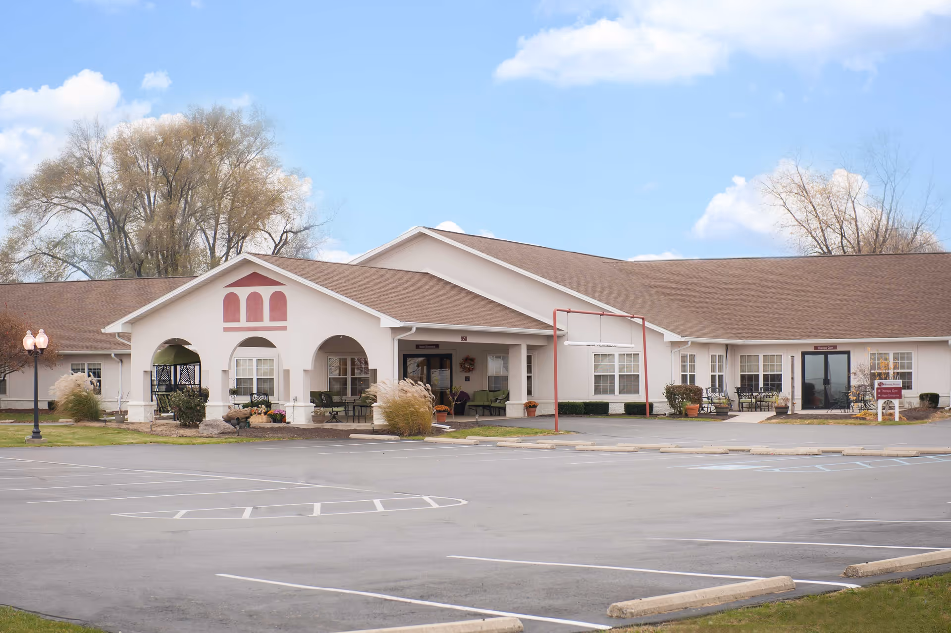 Exterior view of Morning Breeze Retirement Community & Healthcare Center, showing a single-story building with a brown roof, white walls, and a covered entrance with arches. There are outdoor seating areas with chairs and tables, some plants, and a mostly empty parking lot in front. Trees and a partly cloudy sky are visible in the background.