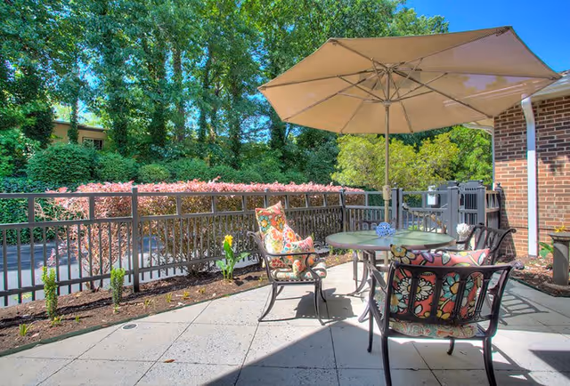 Outdoor patio area with a round table and four metal chairs with colorful floral cushions. A large beige umbrella provides shade over the table. The patio is paved with tiles and bordered by a black metal fence. Behind the fence, there are bushes and tall trees under a clear blue sky.