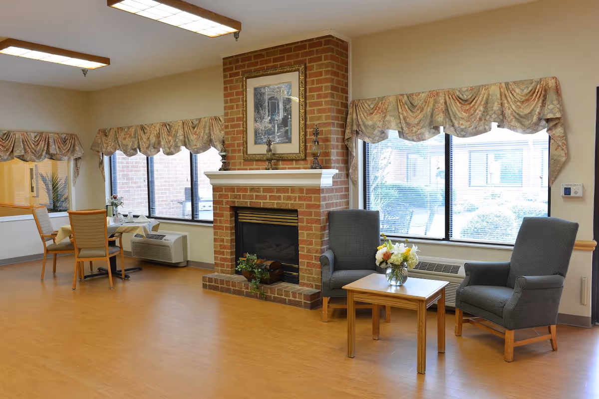 A cozy common area in a senior living facility featuring a brick fireplace with decorative items on the mantel and a framed picture above it. Two gray armchairs are positioned around a wooden coffee table with a vase of fresh flowers. Large windows with floral valances let in natural light, and a small dining table with chairs is visible in the background.
