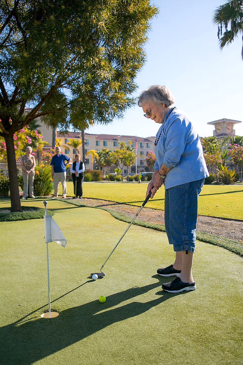 An older woman putting on a small golf green at a senior living community with other residents and buildings in the background.