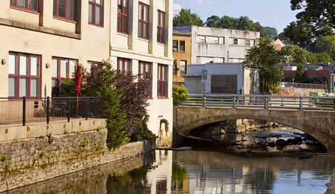Brick riverside building with multiple windows and a small arched bridge crossing a calm river, reflected in the water.