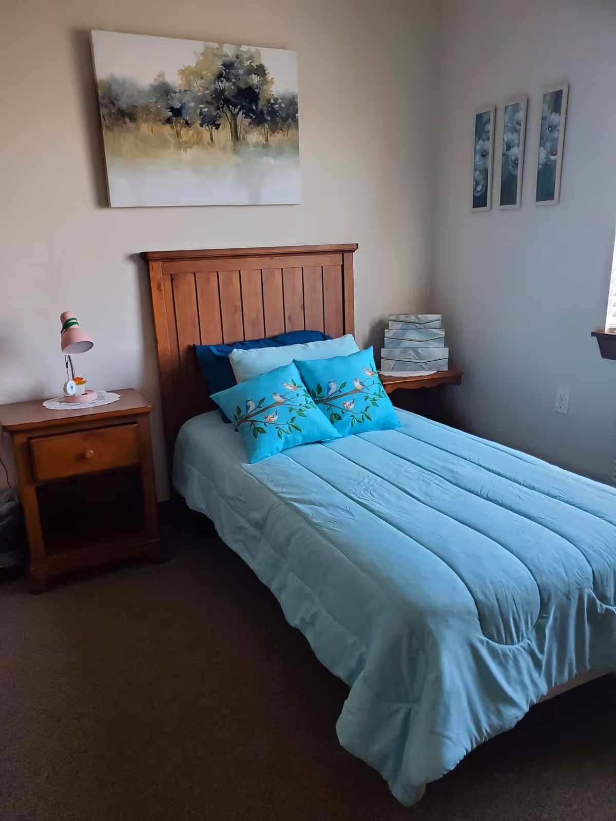 A bedroom with a wooden headboard bed dressed in light blue bedding and two decorative pillows with bird and branch designs. There is a wooden nightstand with a pink lamp and a small clock on the left side of the bed. Above the bed hangs a painting of trees, and on the adjacent wall are three framed floral artworks. A window with blinds is partially visible on the right side.