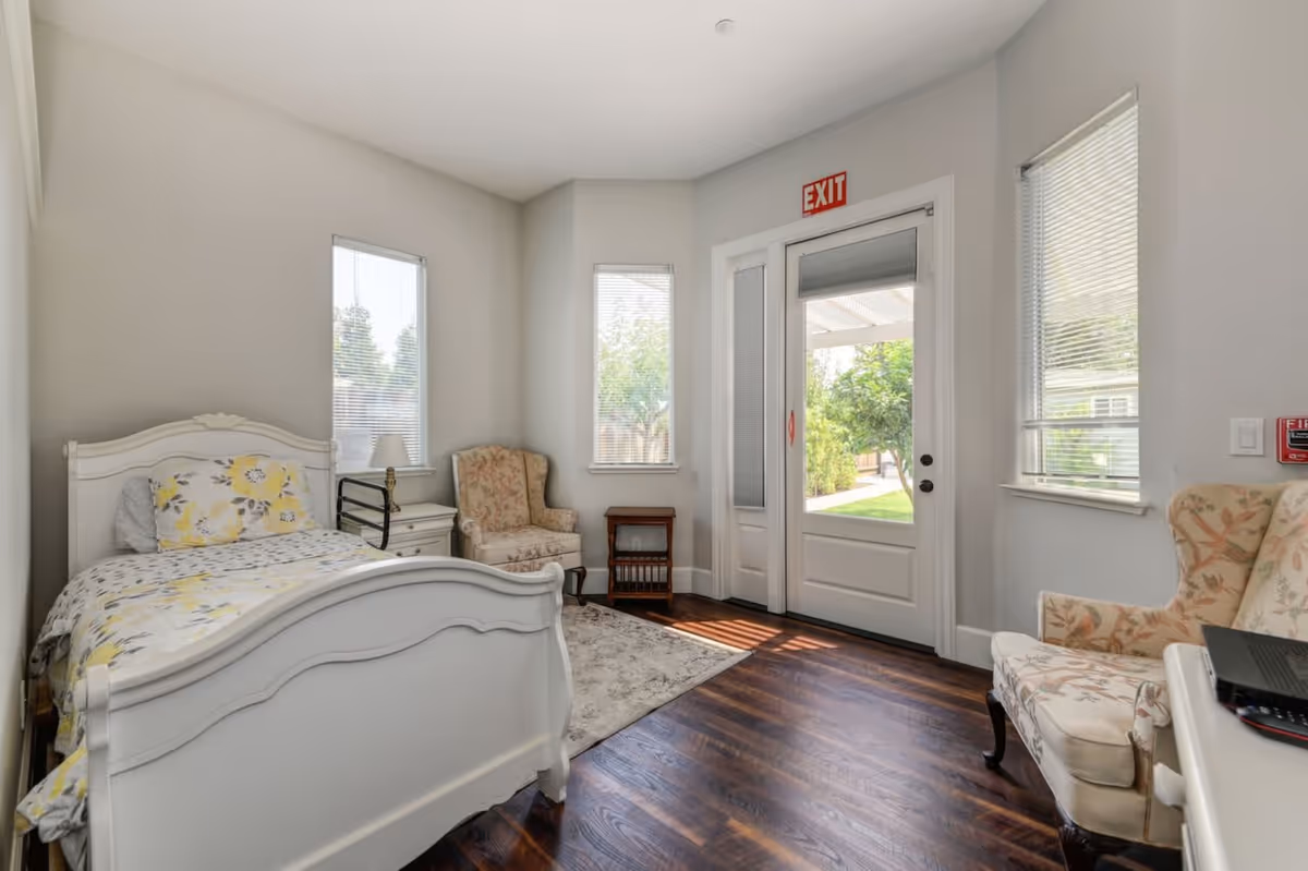 A bright bedroom with a white bed featuring yellow floral bedding, two floral upholstered armchairs, a small white nightstand with a lamp, a small wooden side table, and a door with windows leading outside. The room has wooden flooring, light gray walls, and three windows with blinds. An exit sign is above the door.