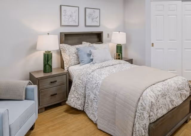 A neatly made bed with floral patterned bedding and multiple pillows in a senior living community bedroom. There are two wooden nightstands on either side of the bed, each with a green lamp. A light blue armchair is partially visible on the left side, and two framed floral artworks hang on the wall above the bed. The room has light-colored walls and wooden flooring.