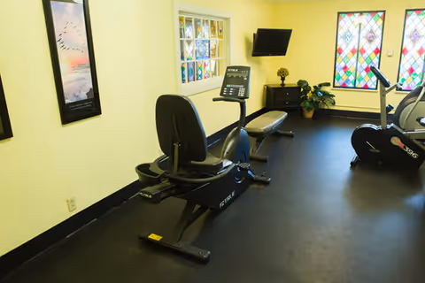 A small exercise room with two stationary bikes and a bench. The room has light yellow walls, a black floor, a wall-mounted TV, a small plant on a cabinet, and colorful stained glass windows.