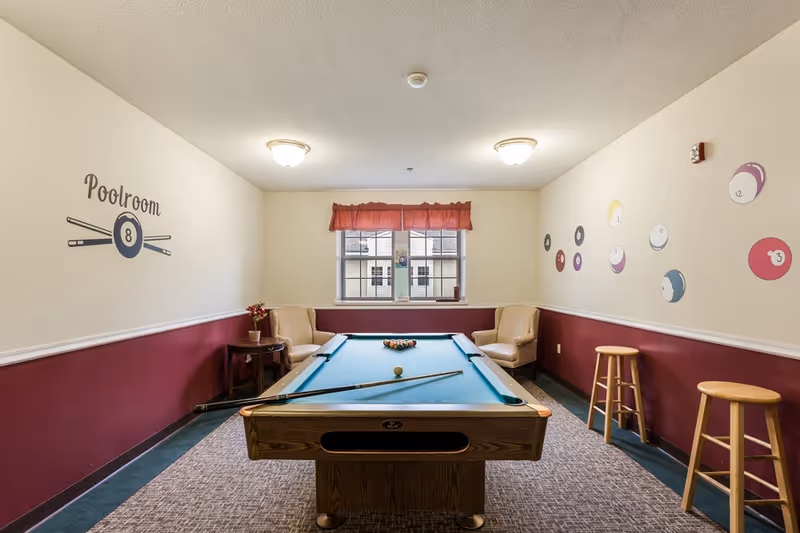 A well-lit pool room with a billiards table centered, two armchairs by a window, stools, and decorative pool ball wall decals.