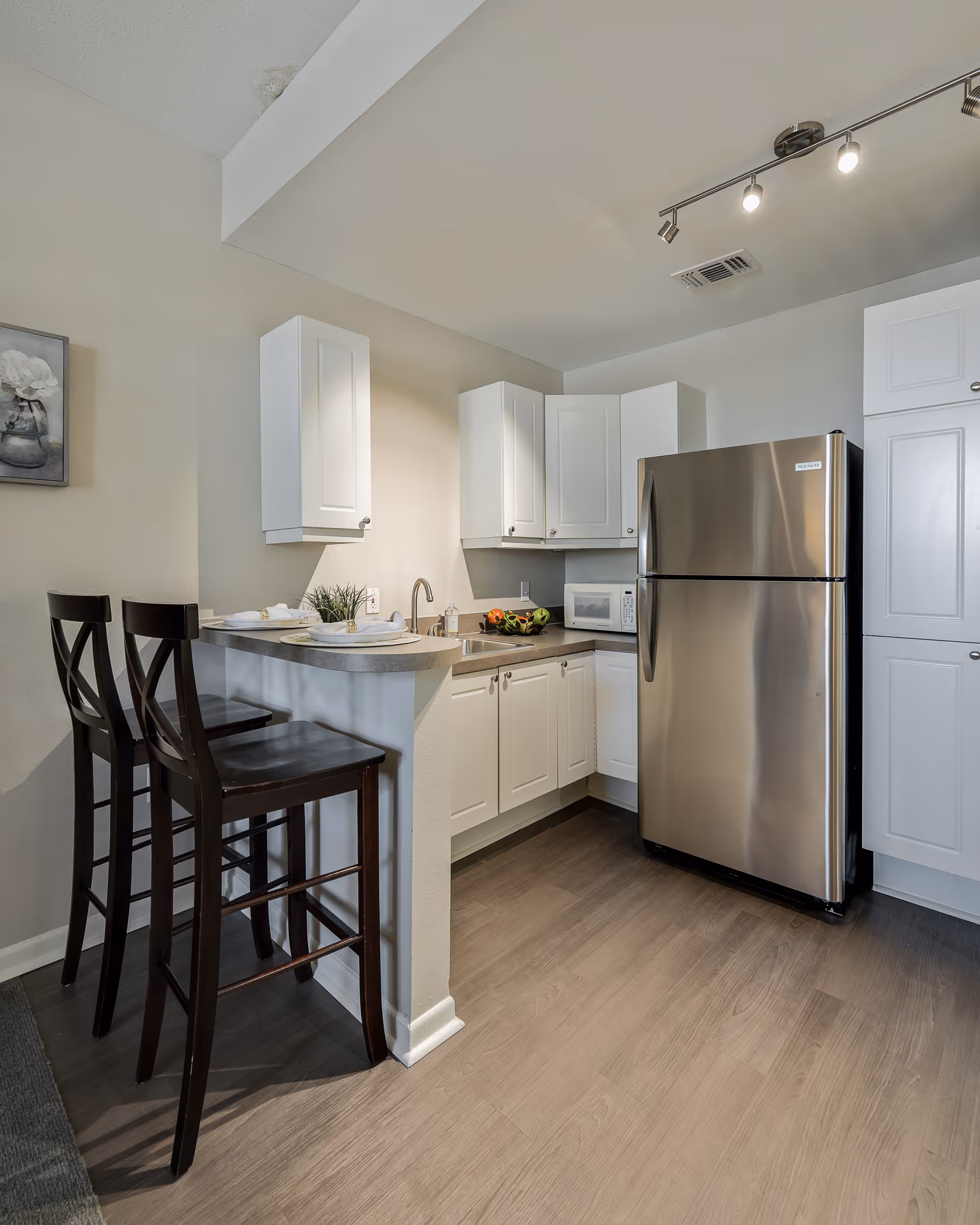 A modern kitchen with white cabinets, a stainless steel refrigerator, a microwave, and a small counter with two dark wooden bar stools. The kitchen has light-colored walls and wood flooring, with a small plant and plates set on the counter.