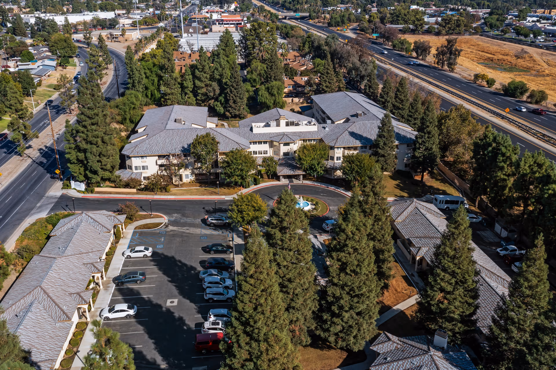 Aerial view of Columbus Estates, a senior living community, showing multiple buildings surrounded by trees, a parking lot with cars, and a circular driveway with a fountain in the center. The community is bordered by roads and highways with visible traffic.