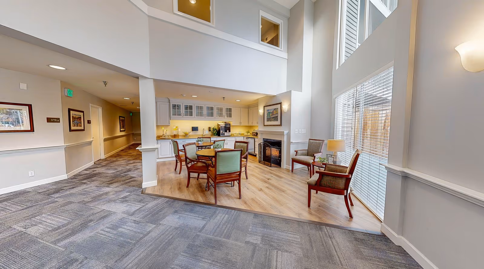 Bright open common area with a round dining table and chairs, kitchenette, fireplace and seating by large windows.