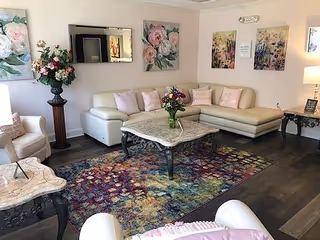 A cozy living room with a white sectional sofa adorned with pastel pink pillows, a colorful patterned rug, and two marble-top coffee tables with dark ornate legs. The walls are decorated with floral paintings and a rectangular mirror. There is a tall vase with flowers on a pedestal and a side table with a lamp and a small framed sign.
