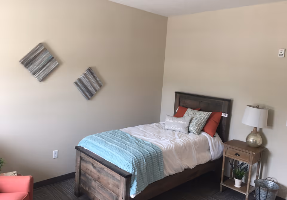 A cozy bedroom with a wooden bed frame, white bedding, and a light blue throw blanket. There are several pillows on the bed, including one with the text 'Home is where the Heart is.' Next to the bed is a wooden nightstand with a lamp and a small potted plant. Two square decorative wall hangings are mounted on the beige wall above the bed. A coral-colored chair is partially visible in the corner.
