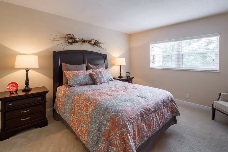 A cozy bedroom with a large bed featuring a patterned bedspread in shades of orange, gray, and white. The bed has multiple pillows and a dark wooden headboard. On either side of the bed are matching dark wooden nightstands with lamps. A window with white blinds lets in natural light, and a decorative wall piece hangs above the headboard. A chair is positioned near the window.