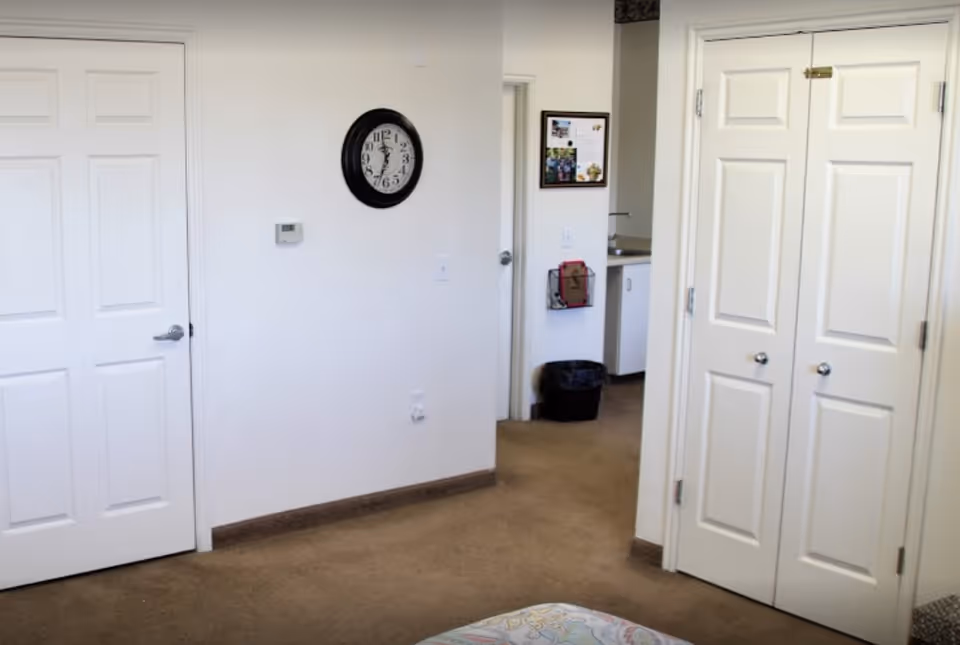 Interior view of a room with beige carpet and white walls featuring two closed white doors and a double-door closet. A black round wall clock is mounted on the wall, and a small hallway leads to a kitchenette area with a sink and a black trash bin underneath. A framed bulletin board is visible on the wall near the kitchenette.