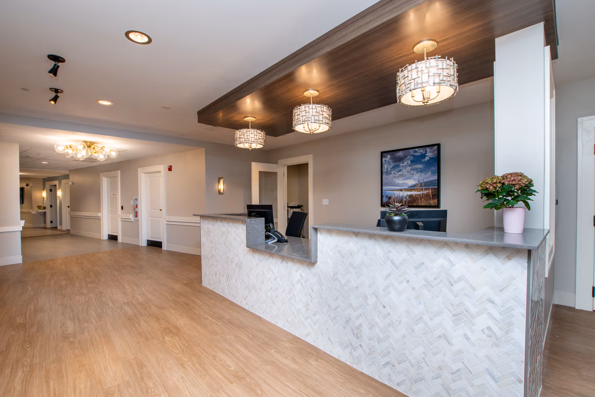 Bright reception desk and lobby area with pendant lights, wood-look flooring, and a framed landscape print on the wall.