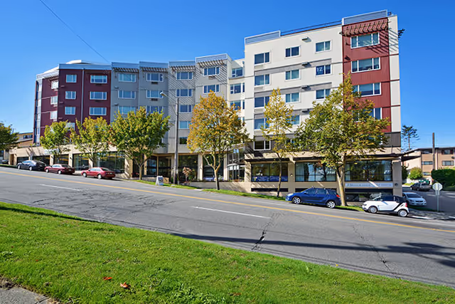 Exterior view of a multi-story senior living facility building with a modern design, featuring red and white sections, several windows, trees along the sidewalk, and parked cars on the street in front.