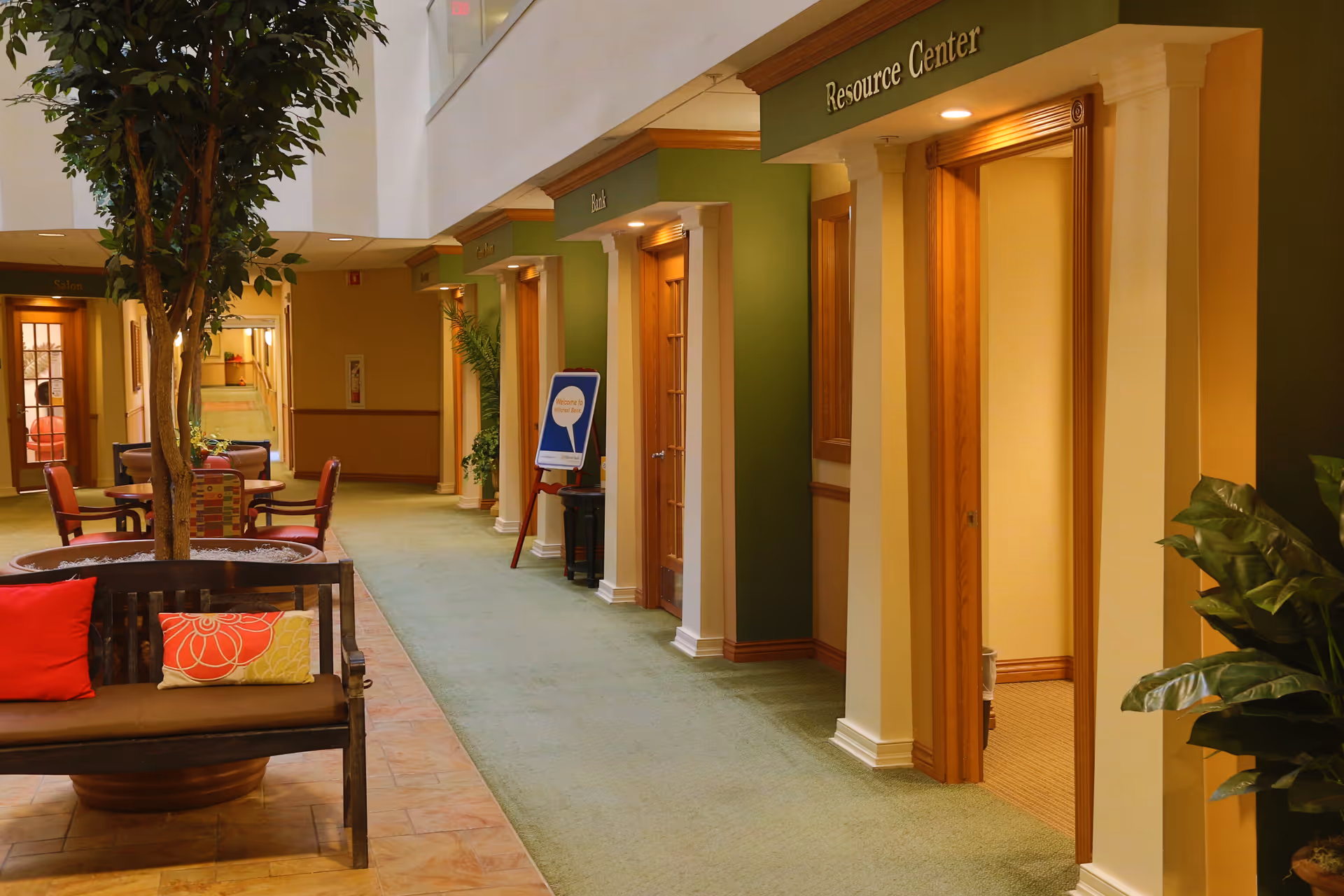Interior hallway of Lakeline Oaks Retirement Community with green carpet and beige walls. Several wooden doors with signs above them, including one labeled Resource Center. A bench with colorful cushions and a large potted tree are visible along the hallway. There are also chairs and tables near the far end of the hallway.