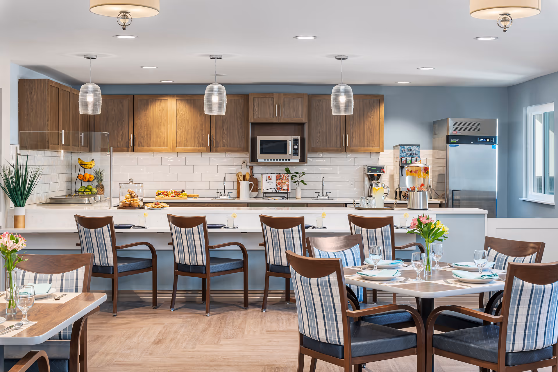 A bright and modern kitchen and dining area in a senior living facility. The kitchen features wooden cabinets, a microwave, a stainless steel refrigerator, and a beverage dispenser. There is a long counter with five wooden chairs with plaid cushions. In the foreground, there are round dining tables set with plates, glasses, and napkins, each decorated with a small vase of flowers. The walls are painted light blue, and the floor has a light wood finish.
