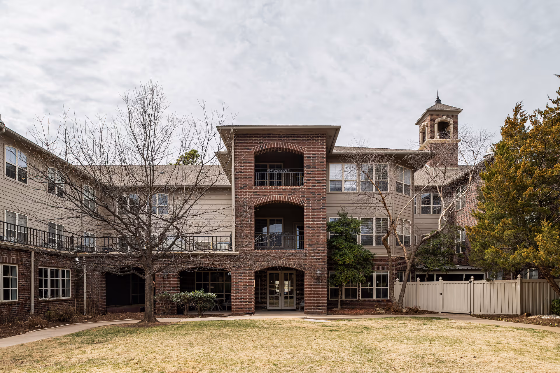 Exterior view of a multi-story assisted living facility building with brick and beige siding, featuring balconies, multiple windows, a bell tower, and a fenced yard with leafless trees and a grassy area in front.