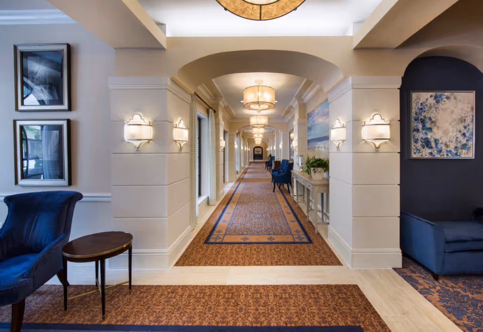 A long, well-lit hallway in a senior living facility with patterned carpet and white walls featuring decorative sconces. Blue upholstered chairs and small tables are placed along the sides, with framed artwork hanging on the walls. The ceiling has multiple round light fixtures, and there is a console table with plants on the right side.