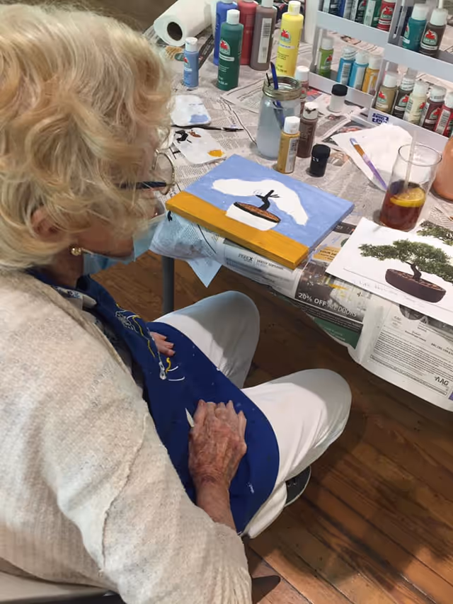 An elderly woman with light blonde hair wearing a face mask and a blue apron is seated at a table covered with newspapers and various bottles of paint. She is painting a picture of a bonsai tree on a canvas. There is a glass of iced tea with a lemon slice on the table, along with a printed reference image of a bonsai tree.