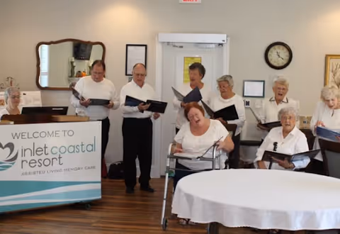 A group of elderly people standing and sitting in a room, some holding songbooks, appearing to sing or perform together. A sign in front reads 'Welcome to Inlet Coastal Resort Assisted Living Memory Care'. The room has wooden floors, a round table with a white tablecloth, a wall clock, and a mirror.