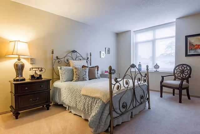 A well-lit bedroom featuring a metal-framed bed with decorative pillows and a light blue patterned bedspread. Next to the bed is a dark wooden nightstand with a lamp and a vintage-style telephone. A window with blinds allows natural light into the room, and a wooden chair with a patterned cushion is placed near the window. The walls are painted light beige, and there is a framed artwork hanging on the wall.