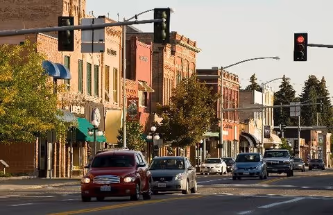 Downtown street lined with brick storefronts, trees and cars stopped at a traffic light.