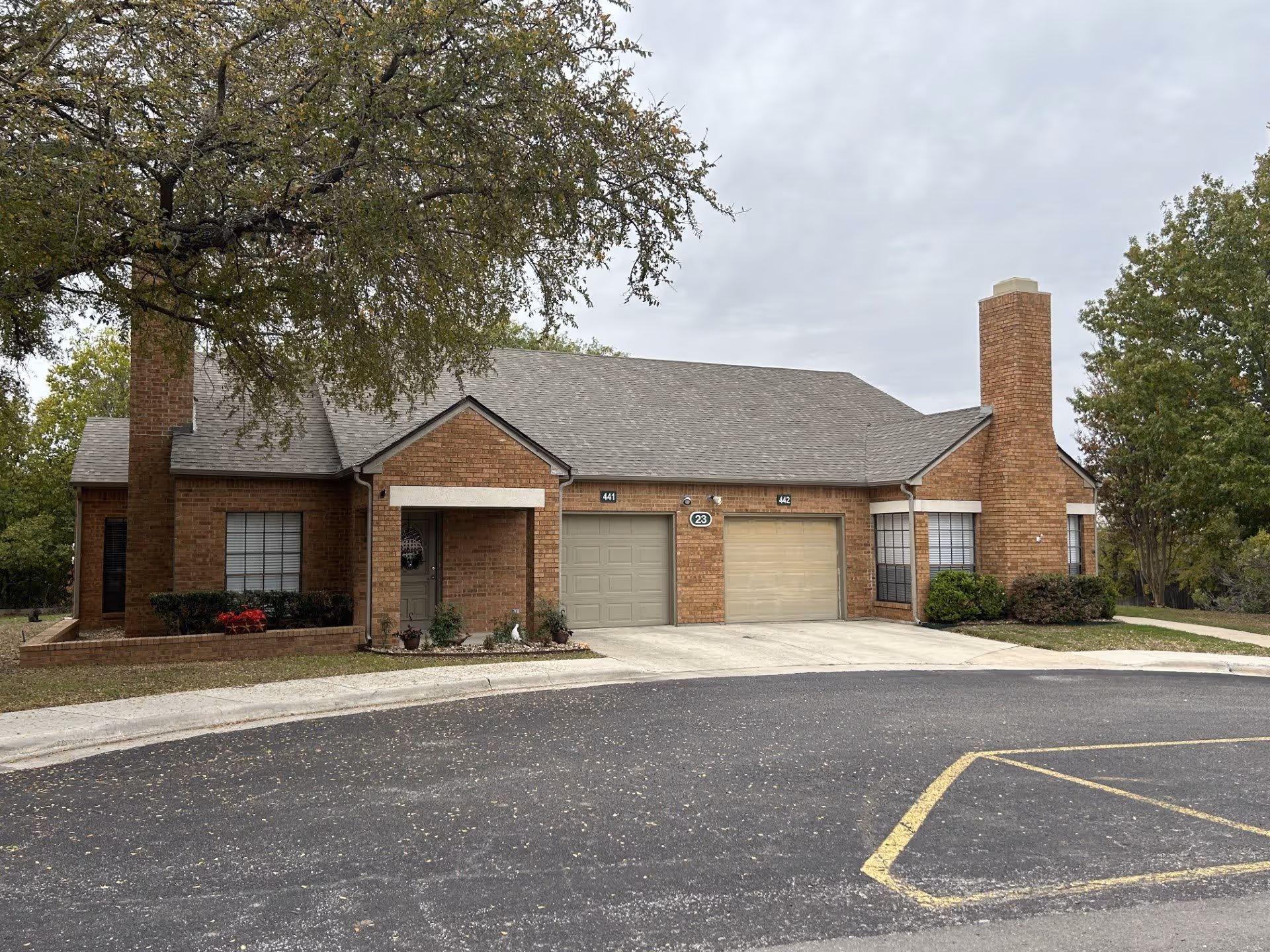 Exterior view of a single-story brick building with two garages and two chimneys, surrounded by trees and a paved driveway with yellow parking markings.