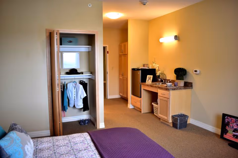 Interior view of a senior living facility room at Sanctuary Grande showing a bed with patterned bedding and pillows in the foreground, an open closet with clothes hanging and shoes on the floor, a small kitchenette area with a mini fridge, sink, countertop, cabinets, and a trash bin, all under warm lighting.