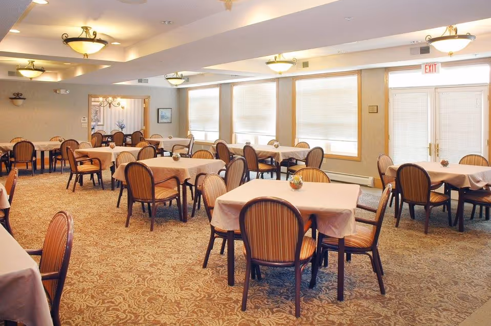Bright dining room with multiple cloth-covered tables and striped chairs, carpeted floor and large windows.
