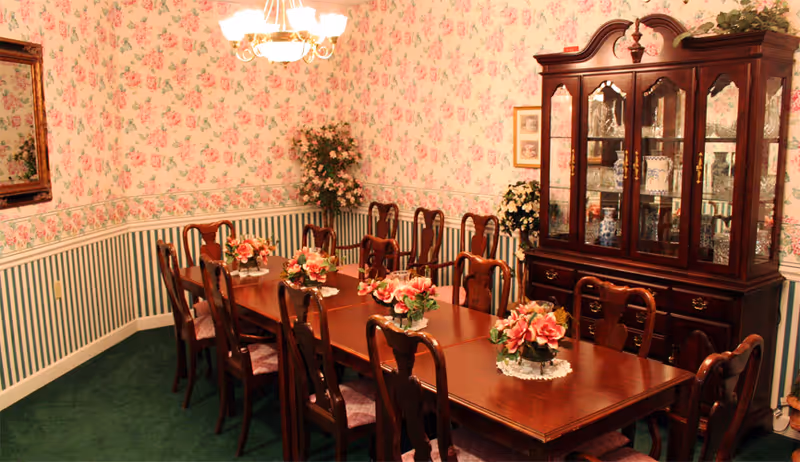 A traditional dining room with a long wooden table surrounded by wooden chairs with cushioned seats. The table is decorated with several floral centerpieces. The walls have floral wallpaper on the upper half and green and white vertical stripes on the lower half, separated by a chair rail. A wooden china cabinet with glass doors displaying dishes and decorative items is against one wall. A chandelier with multiple lights hangs above the table, and there are plants in the corners of the room.