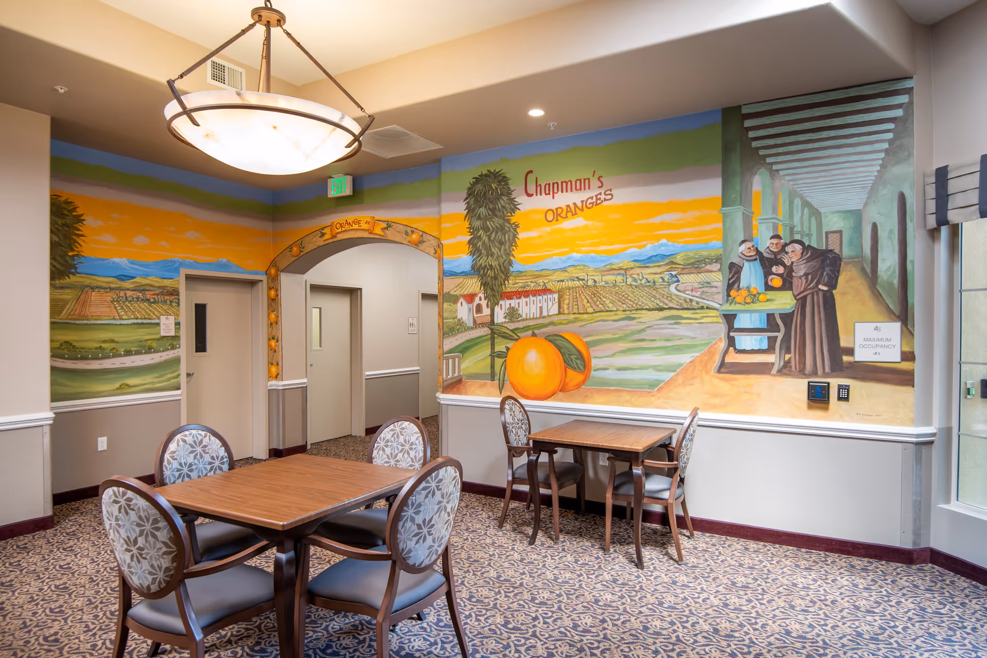 Interior of a senior living facility dining area with two wooden tables and eight upholstered chairs. The walls feature a colorful mural depicting an orange orchard with the text 'Chapman's ORANGES' and three monks examining oranges. There is a large hanging light fixture, patterned carpet, and two doorways in the background.