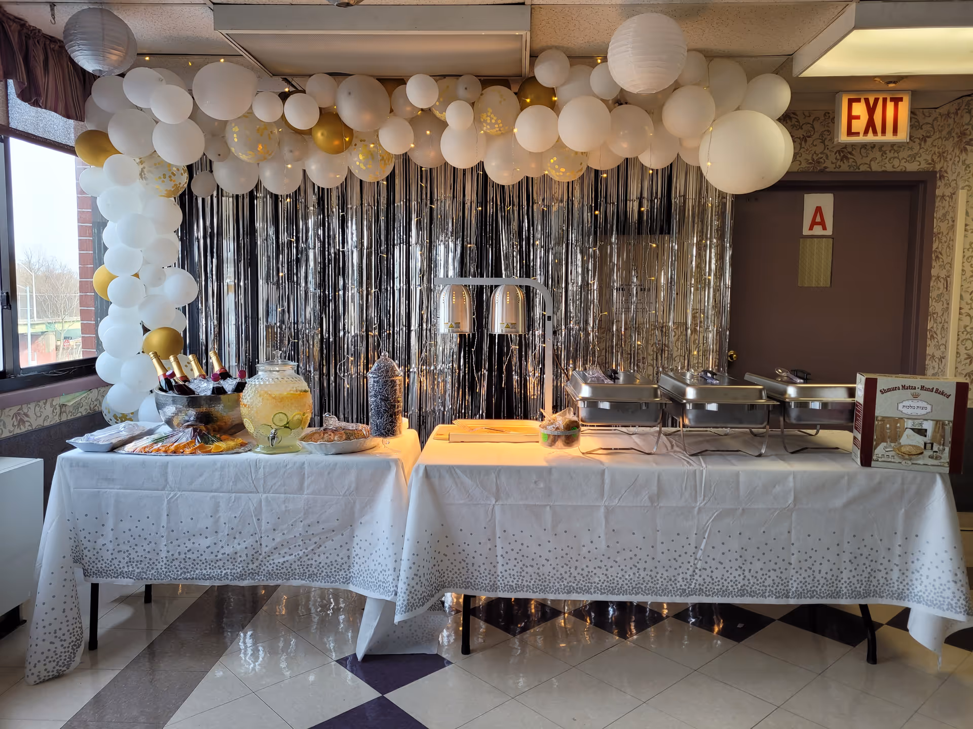Decorated buffet table with white and gold balloons, a silver fringe backdrop, beverage dispenser and chafing dishes set up for a catered event.