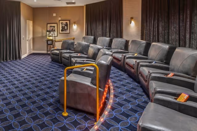 A small private movie theater room with two rows of black leather recliner chairs arranged in a curved formation. The floor is carpeted with a blue and black geometric pattern. There is a yellow handrail with lights along the edge of the step between rows. The walls are beige with dark curtains covering the windows, and framed pictures hang on the far wall.