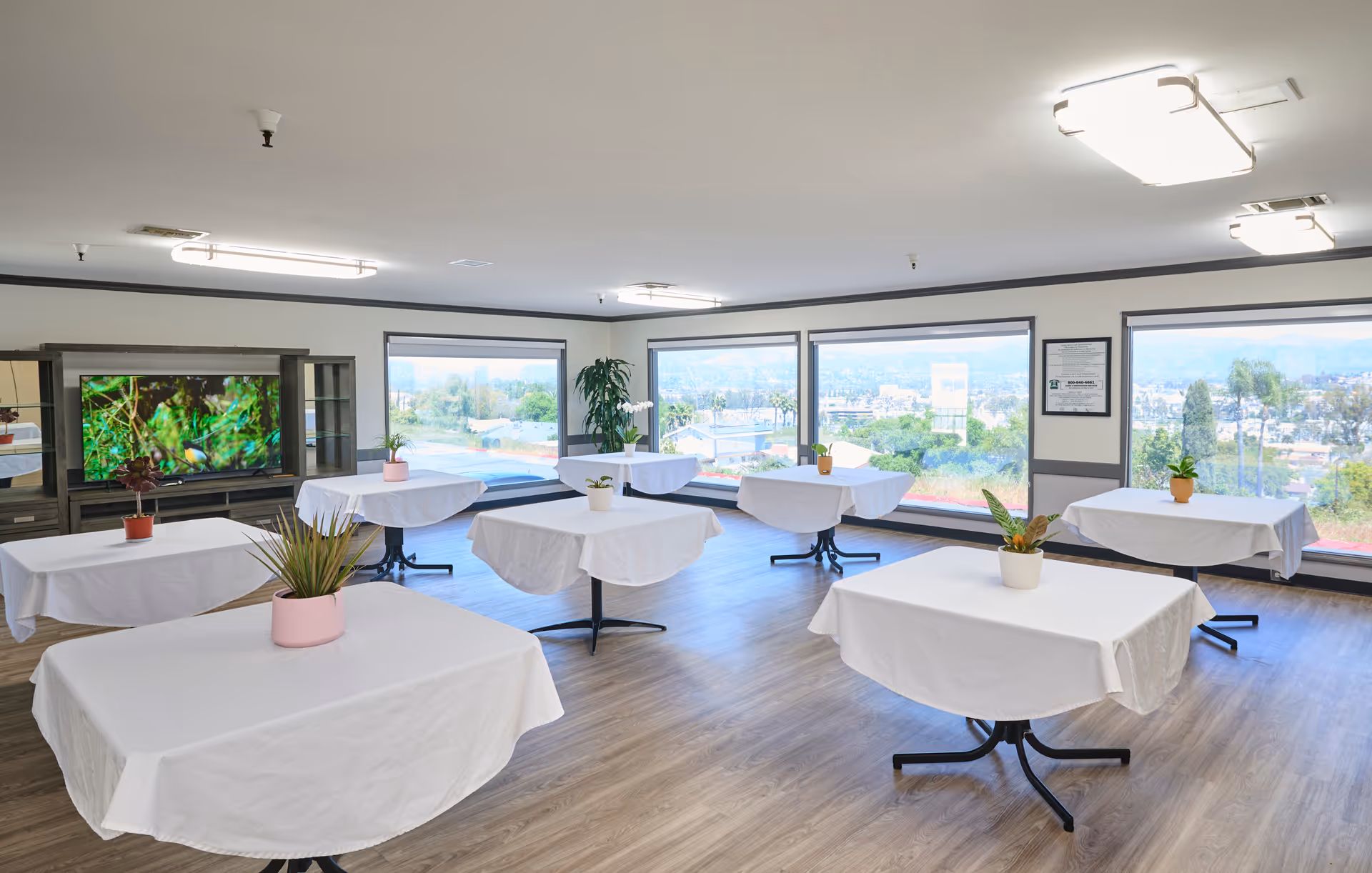 Bright dining/activity room with round tables covered in white cloths and small potted plants and large windows showing an outside view.