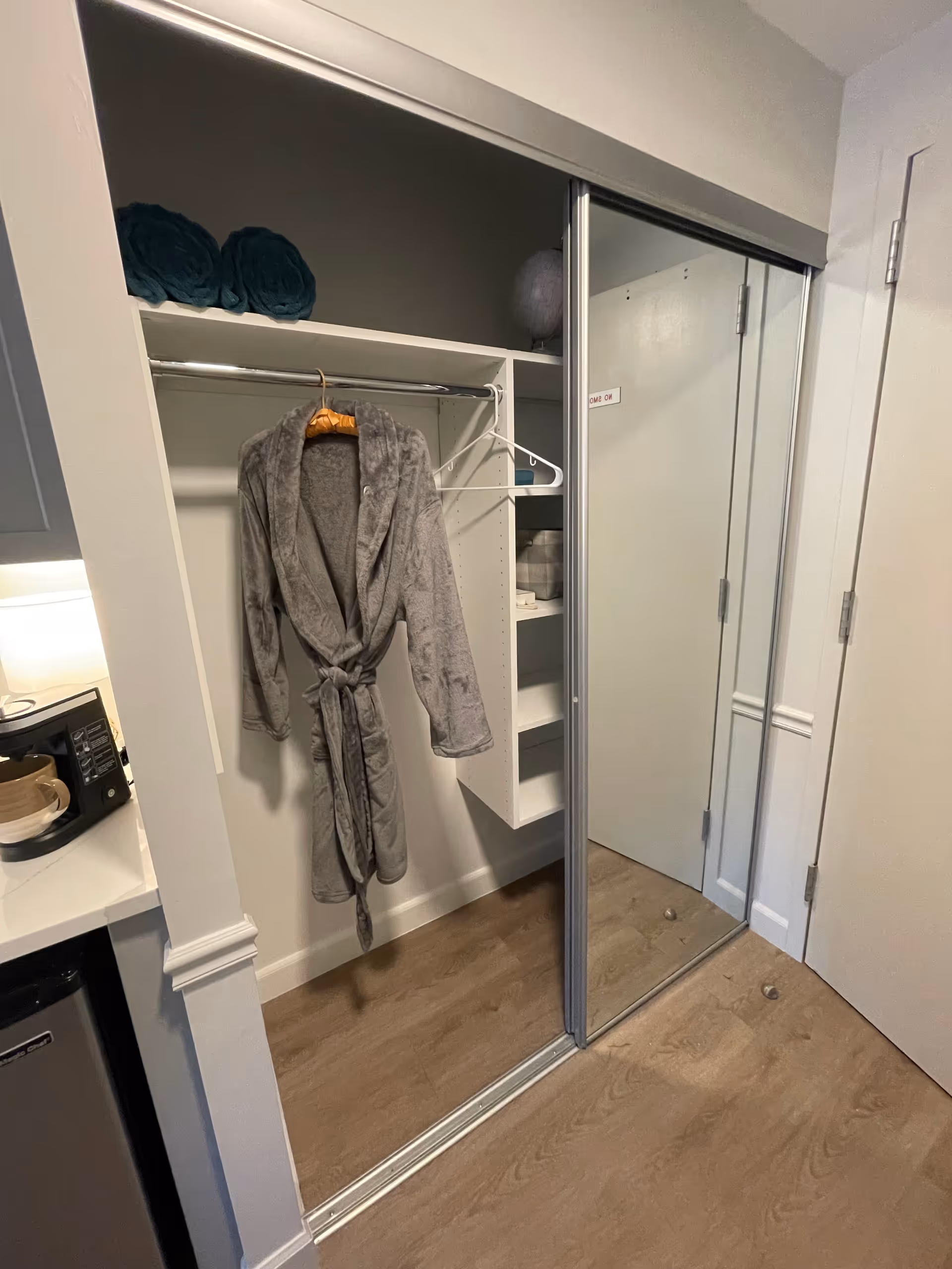 An open closet with sliding mirrored doors in a senior living facility room. Inside the closet, a gray bathrobe hangs on a wooden hanger, and there are two rolled teal towels on the top shelf. To the right, there are built-in shelves with some items placed on them. The floor is wooden, and part of a kitchenette with a microwave and mini fridge is visible on the left side.
