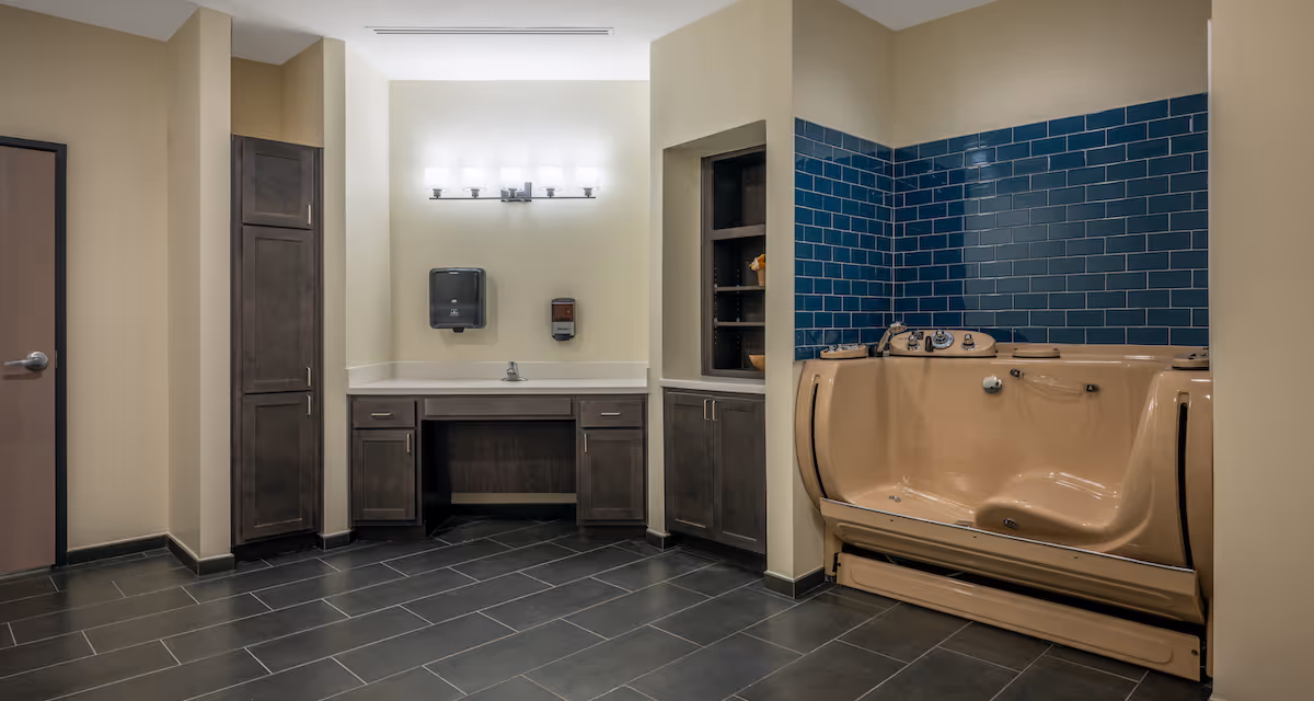 Accessible bathing room with a beige walk-in tub against a dark blue tiled wall, sink vanity and dark cabinetry on a tiled floor.