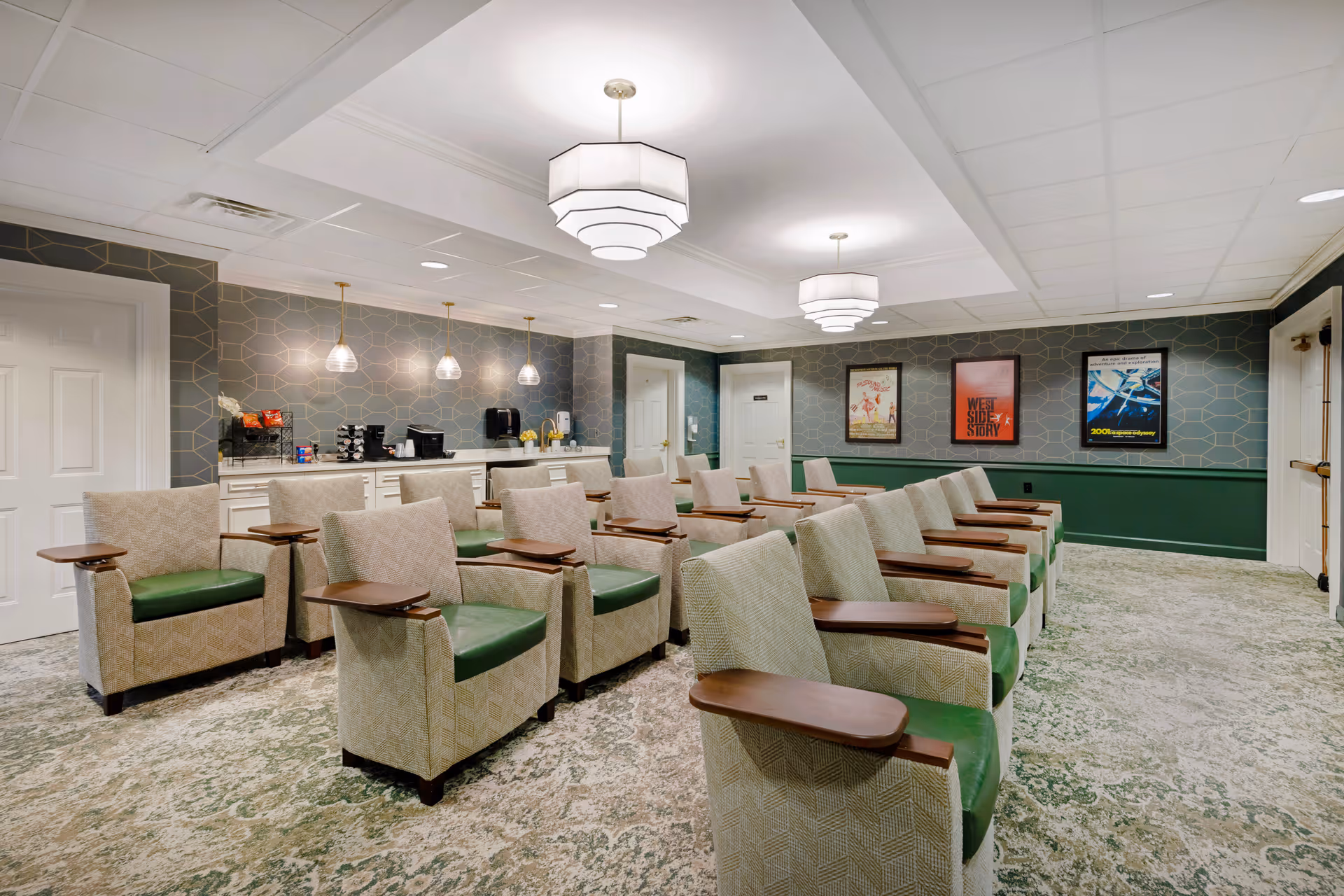 Well-lit communal seating room with rows of upholstered armchairs with green seats and wooden arm trays, pendant lights, and a service counter along the back wall.
