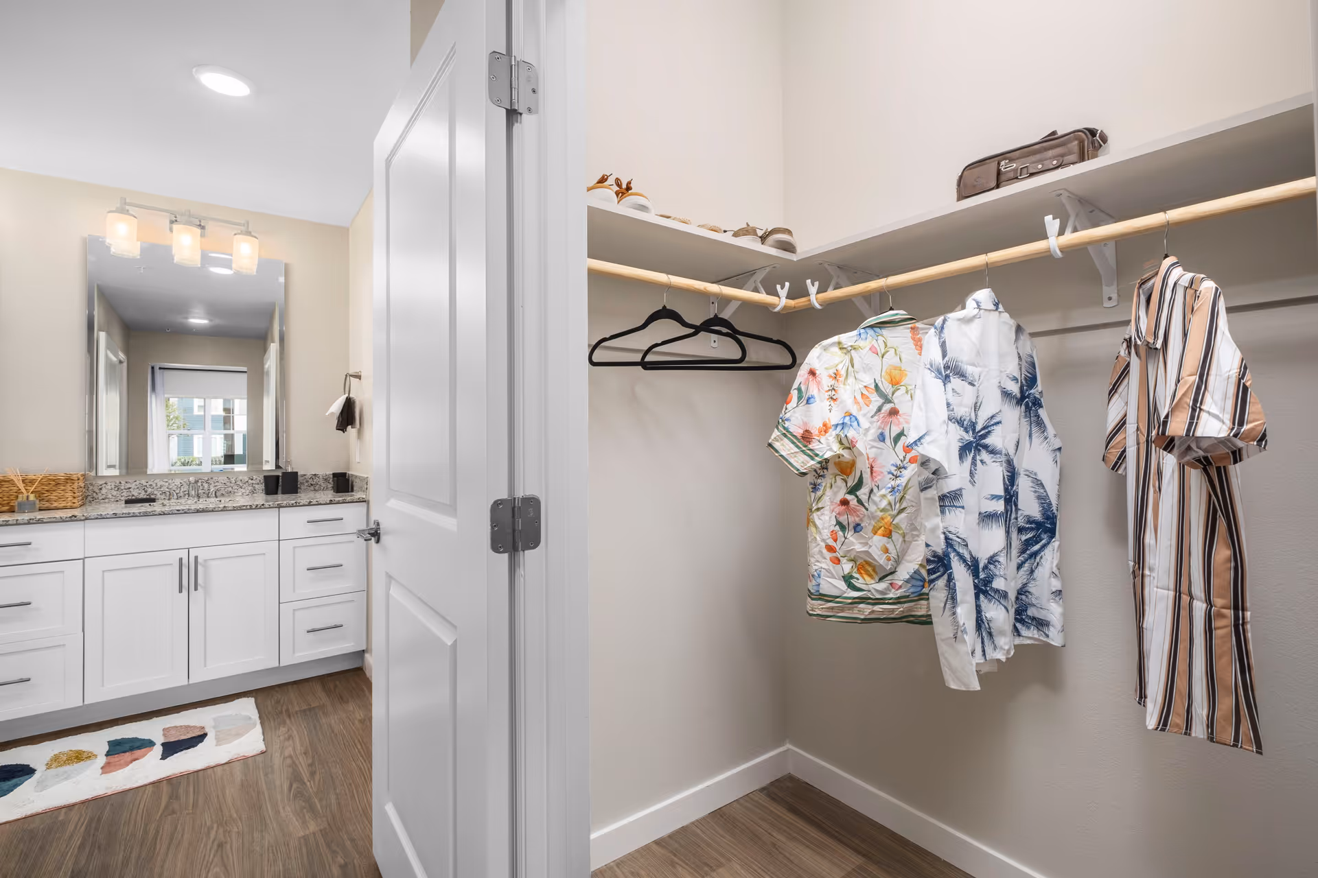 View of a walk-in closet with three hanging shirts and several pairs of shoes on the top shelf, adjacent to a bathroom with a white vanity, granite countertop, mirror, and light fixtures above.