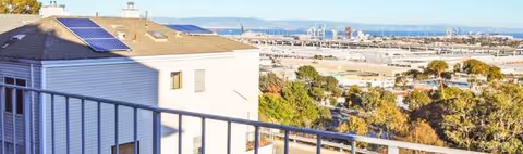 Balcony railing frames a view of rooftops, trees, and a distant cityscape.