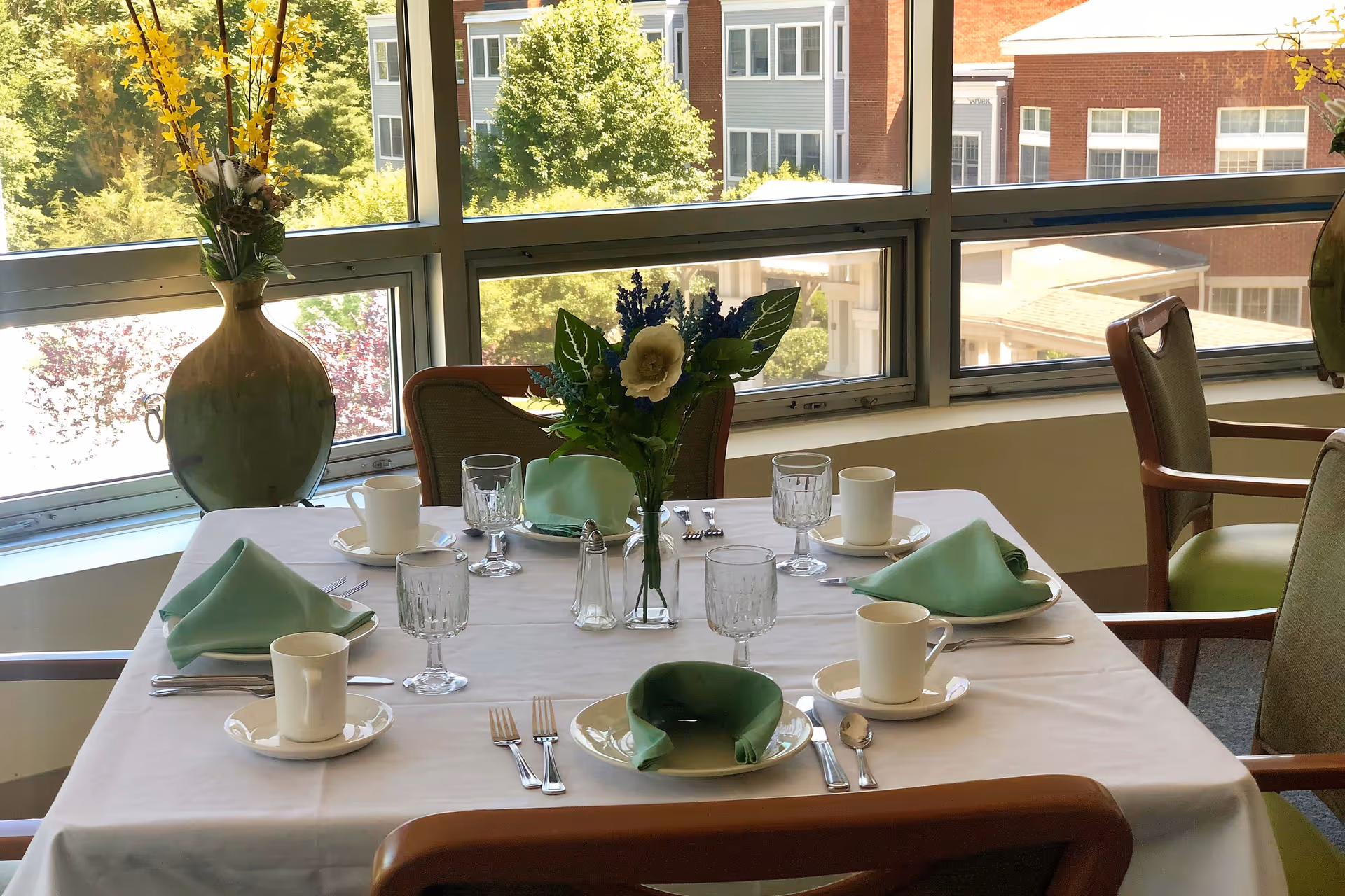 A dining table set for four with white tablecloth, green folded napkins, white cups and saucers, clear water glasses, silverware, and a vase with flowers in the center. The table is near large windows showing trees and a brick building outside.
