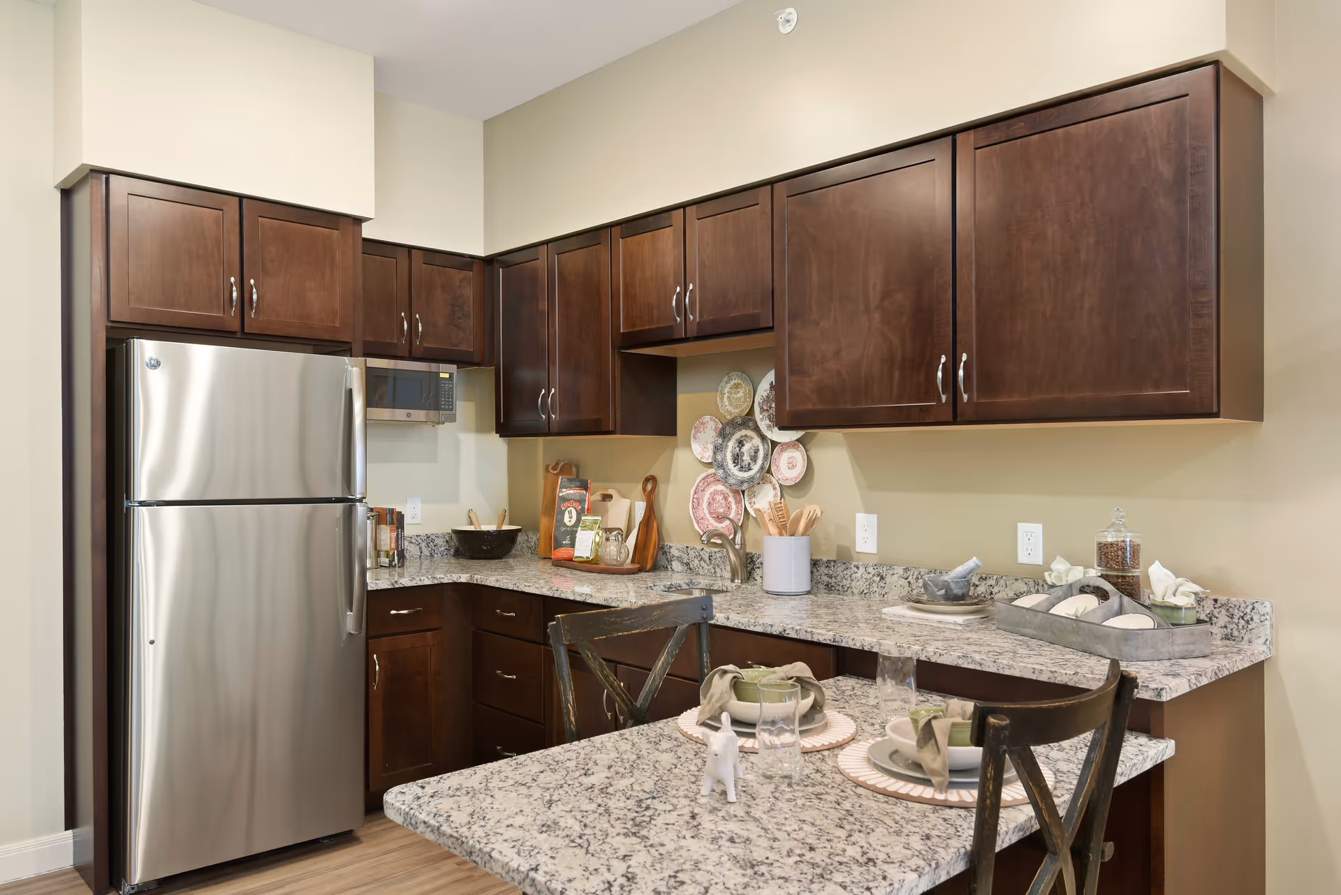 A modern kitchen with dark wooden cabinets, a stainless steel refrigerator, a microwave, and granite countertops. The countertop extends into a small dining area set with plates, bowls, glasses, and napkins. Decorative plates are displayed on the wall above the sink area.
